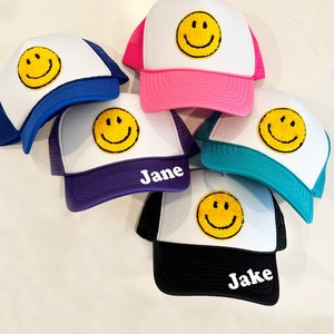 May include: Five colorful trucker hats with a smiling face graphic and personalized names. The hats are white with a black mesh back and feature different colored brims. The names on the hats are "Jane", "Jake", and three other names that are not visible in the image.