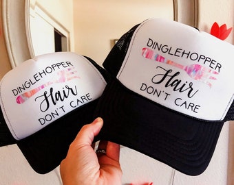 Puede incluir: Dos gorras de camionero en blanco y negro con el texto "Dinglehopper Hair Don't Care". Las gorras tienen un panel frontal blanco con un gráfico de tenedor colorido. Las gorras están sostenidas por una mano.