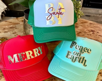 May include: Three Christmas themed trucker hats. The first hat is white with green mesh and says "Baby Santa" in gold glitter letters. The second hat is red with black mesh and says "Merry" in gold glitter letters. The third hat is light blue with green mesh and says "Peace on Earth" in gold glitter letters.