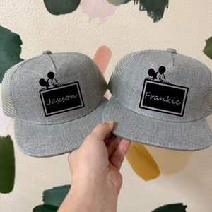 May include: Two gray and black baseball caps with a black and white Mickey Mouse graphic and a black rectangle with a name on it. The names on the caps are Jaxson and Frankie.