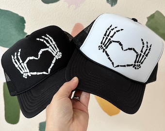 May include: Two black trucker hats with white mesh back panels. The front of each hat features a white skeleton hand design making a heart shape.