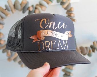 Puede incluir: Una gorra de camionero gris con una malla negra en la parte posterior. La gorra tiene un diseño de lámina dorada que dice "Once Upon A Dream".