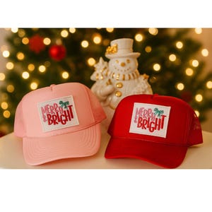 Puede incluir: Dos gorras de camionero, una rosa y otra roja, con la parte trasera de malla y una visera curva. Cada gorra presenta un parche rectangular con las palabras "Merry & Bright" en una fuente festiva, acentuado con un lazo verde. Una figura de muñeco de nieve está en el fondo.