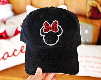 Puede incluir: Gorra de béisbol de pana negra con un contorno blanco y plateado de la cabeza de Mickey Mouse con un lazo rojo y plateado de purpurina.