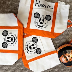 K&ouml;nnte beinhalten: Drei wei&szlig;e Canvas-Tragetaschen mit orangefarbenem Besatz. Jede Tasche zeigt ein Schwarzwei&szlig;bild von Mickey Mouse mit einem K&uuml;rbis-Kopf und Spinnweben. Die oberste Tasche tr&auml;gt den Namen "HARLOW" aufgedruckt.