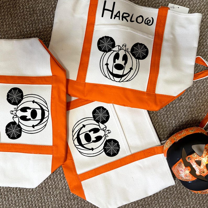 K&ouml;nnte beinhalten: Drei wei&szlig;e Canvas-Tragetaschen mit orangefarbenem Besatz. Jede Tasche zeigt ein Schwarzwei&szlig;bild von Mickey Mouse mit einem K&uuml;rbis-Kopf und Spinnweben. Die oberste Tasche tr&auml;gt den Namen "HARLOW" aufgedruckt.