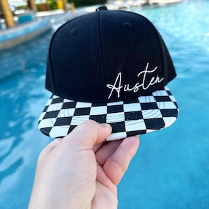 May include: Black baseball cap with a chequered brim and the name "Austen" in white script. The cap is held up against a blue water background.