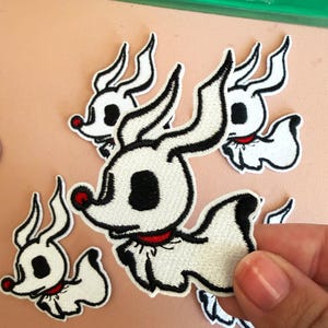 Puede incluir: Un conjunto de parches bordados termoadhesivos que representan un perro fantasma blanco con grandes ojos negros, un collar rojo y una pequeña nariz roja. Los parches tienen un contorno negro cosido distintivo y un cuerpo blanco, en grupo.