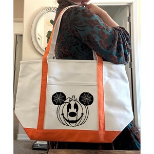 K&ouml;nnte beinhalten: Eine wei&szlig;e Canvas-Tragetasche mit orangefarbenem Besatz zeigt eine schwarz-wei&szlig;e Grafik eines K&uuml;rbisses mit Mickey-Maus-Ohren und einem Spinnennetz-Design.