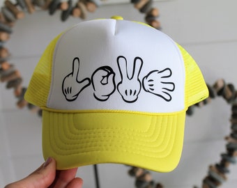 Puede incluir: Una gorra de camionero amarilla y blanca con un diseño negro de manos que deletrean "LOVE" en lenguaje de señas.