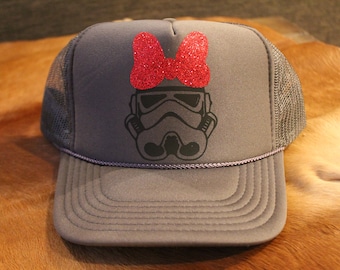 May include: A gray trucker hat with a black and white image of a Stormtrooper helmet and a red glitter bow on top.