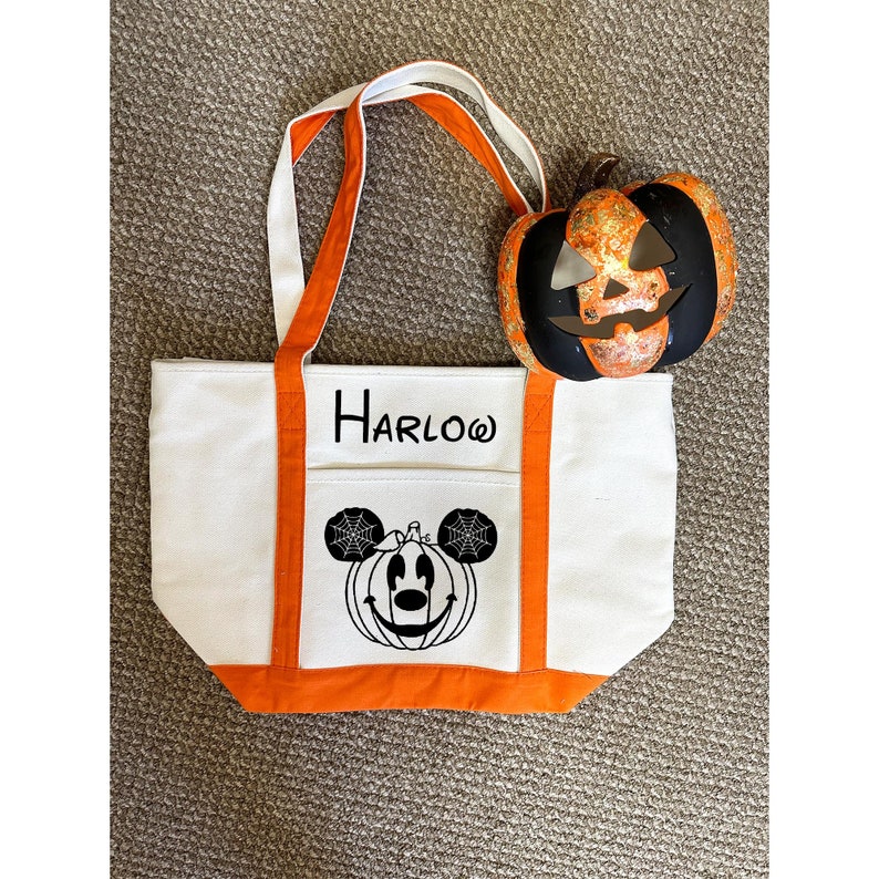 K&ouml;nnte beinhalten: Wei&szlig;e Canvas-Tragetasche mit orangefarbenem Besatz. Die Tasche hat ein schwarz-wei&szlig;es Bild von Mickey Mouse, der als K&uuml;rbis verkleidet ist, mit einem Spinnennetz an den Ohren. Die Tasche tr&auml;gt auch den Namen "HARLOW" auf sich.