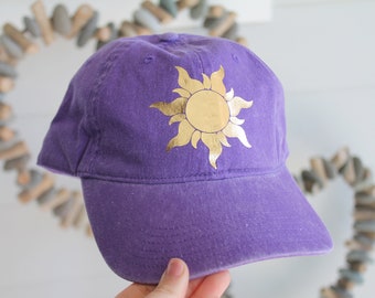 Puede incluir: Gorra de béisbol morada con un diseño de sol dorado.
