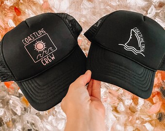 Op de afbeelding: Twee zwarte truckerpetten met mesh ruggen. Eén pet heeft een roze zon- en golfafbeelding met de tekst "COASTLINE CREW". De andere heeft een witte haaienvin-afbeelding en de tekst "COASTLINE CREW".