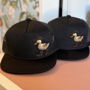 Puede incluir: Dos gorras de camionero negras con la parte trasera de malla y una visera curva. Cada gorra presenta un diseño bordado de un pato con sombrero y botas de vaquero. Las gorras se exhiben sobre una superficie rosa.