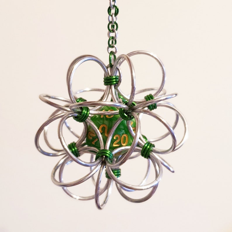 May include: A silver metal cage pendant with a green die inside. The die has gold numbers and is held in place by green wire. The pendant is suspended by a silver chain. The die shows the number 20.