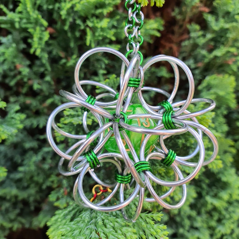 May include: A silver wire ornament with a green die at its core, featuring gold numerals. The wire is accented with green. It hangs from a green chain. The background is a blurred green bush.