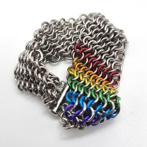 Pode incluir: Uma pulseira de cota de malha prateada com um detalhe arco-íris. A pulseira apresenta uma série de anéis de metal interligados, com uma seção de anéis coloridos em vermelho, laranja, amarelo, verde, azul e roxo. A pulseira tem um fecho prateado.