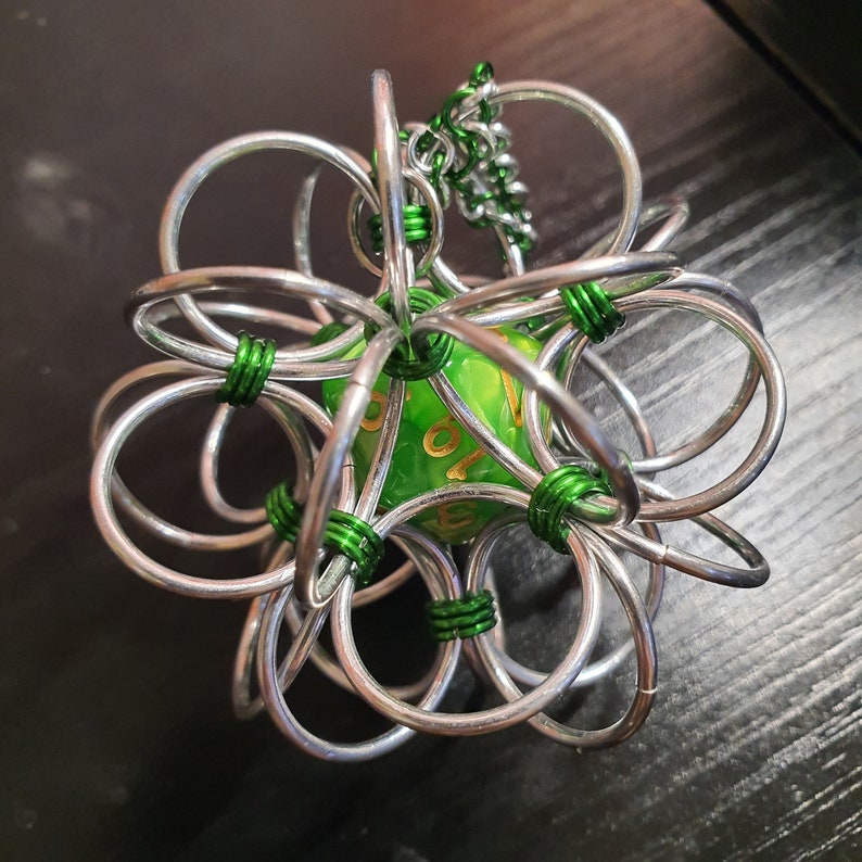 May include: A handcrafted, silver-coloured metal cage encasing a green, multi-sided die. The cage is constructed from interlocking circular rings, with green wire accents. The die has gold-coloured numbers. The item is designed for tabletop gaming.