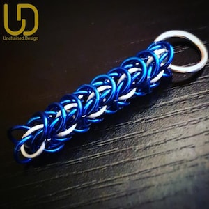 Può includere: Un portachiavi fatto a mano con un design a maglia di catena. Il portachiavi è realizzato con anelli di metallo blu e bianco, collegati a un anello diviso color argento. Il testo "Unchained.Design" è visibile nell'angolo in alto a sinistra.