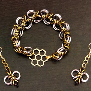Op de afbeelding: Een set sieraden met een zwarte en zilveren kettingarmband met een gouden honingraatbedel, en bijpassende zwarte en zilveren oorhangers met een gouden ketting en honingraatbedel.