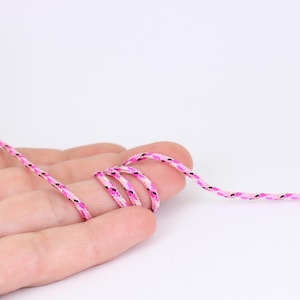 May include: A close-up shot of a pink and white braided cord with black flecks. The cord is looped around a finger, highlighting its flexibility and texture. The cord's design features a repeating pattern of pink, white, and black.