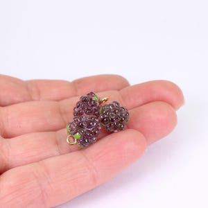 May include: Three small, purple glass charms shaped like blackberries. Each charm has a gold-coloured loop and green accents. The charms are held in a person's hand against a white background. These charms are ideal for jewellery making.