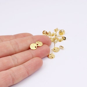 May include: Gold-colored earring posts and backs. The earring posts have a textured, hammered appearance with a round disc shape. The earring backs are butterfly style. Several pieces are scattered on a white surface.