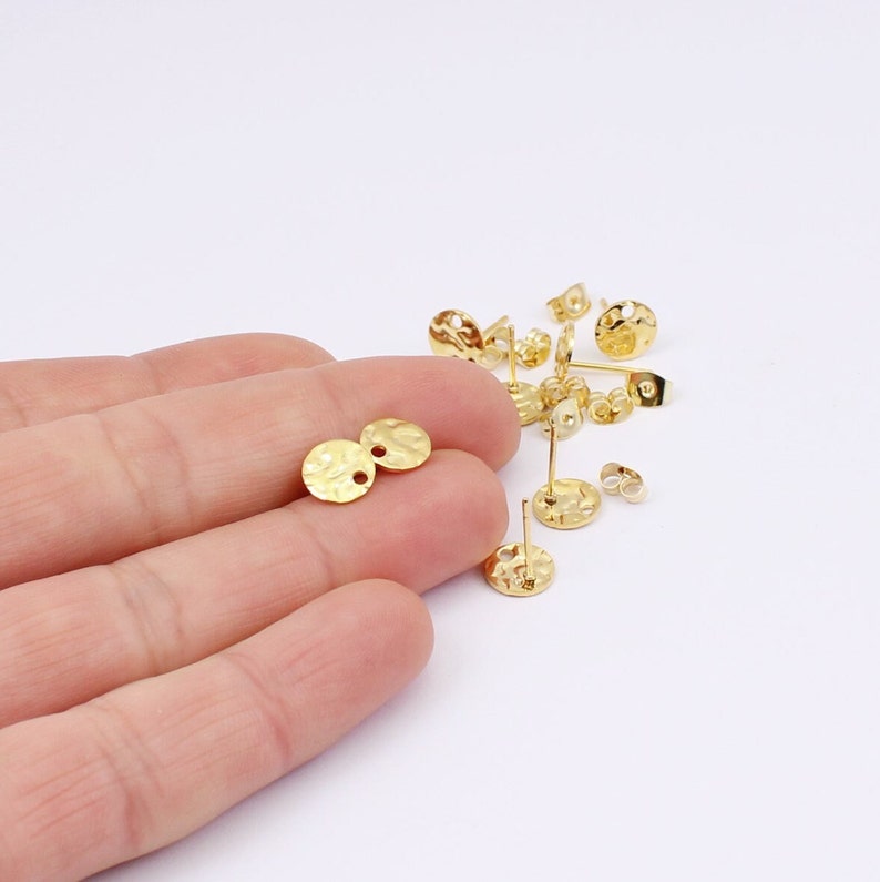 May include: Gold-colored earring posts and backs. The earring posts have a textured, hammered appearance with a round disc shape. The earring backs are butterfly style. Several pieces are scattered on a white surface.