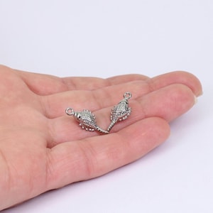 Könnte beinhalten: Zwei kleine, silberfarbene Muschelanhänger. Jeder Anhänger hat eine detaillierte, strukturierte Oberfläche und eine kleine Schlaufe oben zum Befestigen an Schmuck. Die Anhänger werden vor einem neutralen Hintergrund präsentiert.