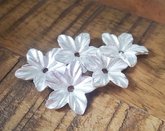 May include: Five silver flower-shaped charms with a hole in the centre. The charms are arranged in a cluster on a wooden surface.