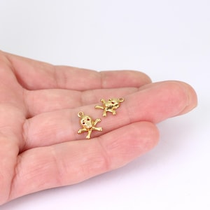 May include: Two small, gold-coloured skull charms with red accents in the eye sockets. The charms have a loop at the top for attaching to jewellery. The skulls have crossed bones below. The charms are displayed on a light background.
