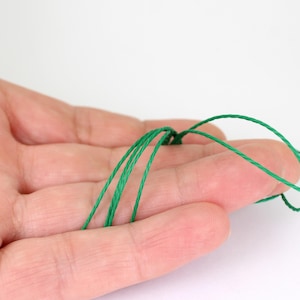 May include: Close-up of a hand holding several strands of bright green thread. The thread is thin and appears to be made of a smooth, possibly synthetic material. The background is plain white, highlighting the thread's color and texture.