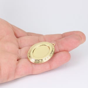May include: A gold-colored oval locket with a decorative border. The locket has a smooth, blank center and a small loop at the top for attaching to a chain. The locket is held in an open hand against a white background.