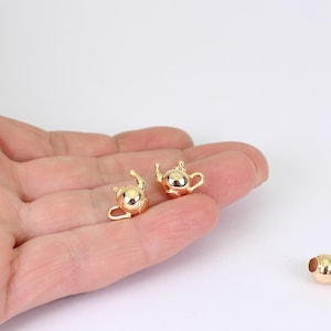 May include: Two small, gold-colored teapot charms. The charms are shiny and detailed, with a spout, handle, and lid. The teapots are likely for jewelry making or crafting. The charms are held in a hand against a white background.
