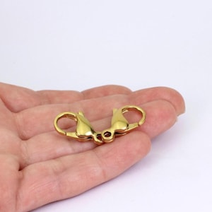 May include: A gold-coloured lobster clasp, with a spring-loaded mechanism for secure closure. The clasp has a rounded shape with a curved lever and a small loop on each end. The metal has a shiny, reflective surface.