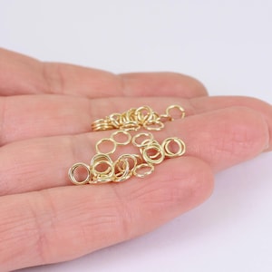 May include: A selection of small, gold-coloured jump rings, scattered across a hand. These circular rings are used for jewellery making and crafting. The rings are made of a shiny, metallic material.