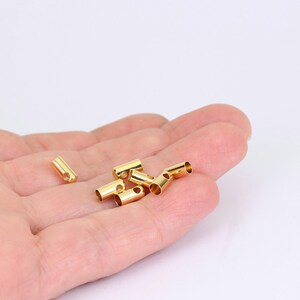 May include: Several small, gold-colored metal tube beads with a hole at one end are displayed. The beads are cylindrical and have a shiny finish. They are scattered on a light background, suggesting they are craft or jewelry-making supplies.