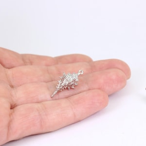 May include: A small, silver seashell charm with a textured surface and a loop at the top for attaching to a necklace or bracelet. The charm is in the shape of a spiky conch shell.