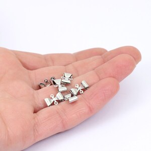 May include: A collection of small, shiny silver-colored metal crimp end findings. These findings have a decorative shape with a ribbed design and are used in jewelry making. The findings are shown against a white background.