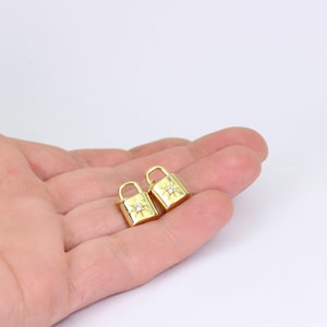 May include: Two small, gold-colored padlock charms with a starburst design and a small, clear gem in the center. The charms are held in a person's hand against a white background. The charms are likely for jewelry making.