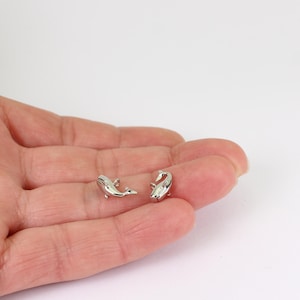 May include: A pair of small, silver whale earrings. The earrings are shaped like whales with fins and tails. The earrings are shown on a person's hand.