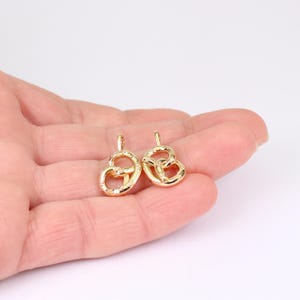 May include: Two gold-colored pretzel-shaped charms. Each charm has a small loop at the top for hanging. The pretzels have a textured surface and are displayed against a white background.