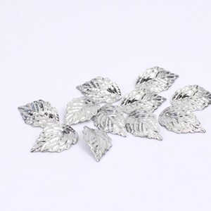 May include: A group of silver metal leaves, scattered on a white surface. The leaves have a textured, slightly raised design.
