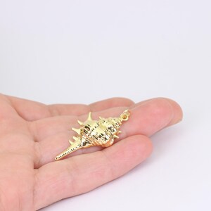 May include: A gold-coloured seashell pendant with a textured surface and a small loop at the top for attaching to a chain or cord. The pendant is approximately 5 cm long and has several pointed protrusions.