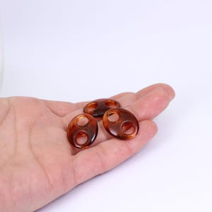 以下が含まれることがあります： 中央に丸い穴が開いた、茶色の楕円形の樹脂ビーズが3つ。ビーズはべっ甲模様で、明るい背景に表示されています。ジュエリー作りに適しています。