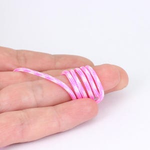 May include: A coiled pink and white cord, possibly a shoelace, is held in a hand. The cord is tightly wound, displaying its pattern and texture. The background is plain white.