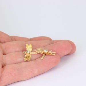 Könnte beinhalten: Zwei goldfarbene Libellen-Anhänger mit filigranen Flügeln und einem klaren Kristallakzent auf dem Körper. Die Anhänger werden vor einem hellen Hintergrund präsentiert, der ihr filigranes Design hervorhebt.