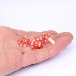 May include: Three red and white polka dot mushroom charms with gold accents. The charms are on a hand.