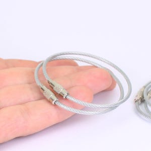 以下が含まれることがあります： 編み込みスチールケーブルデザインのシルバーカラーのキーリング2つ。各リングにはネジ式のコネクタが付いています。リングは白い背景に表示されています。